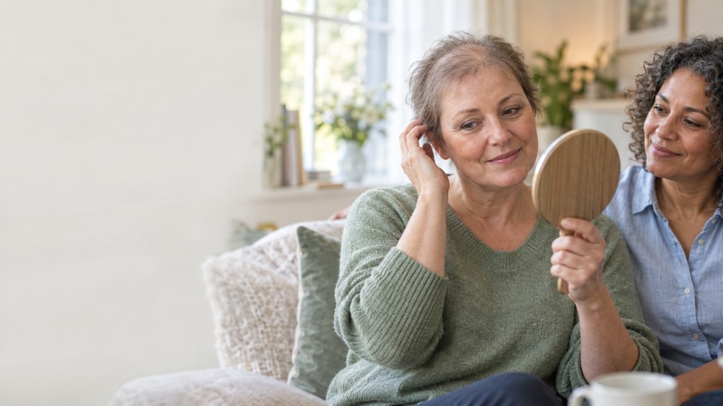 Woman with thinning hair looking in a mirror while receiving support from a friend at home