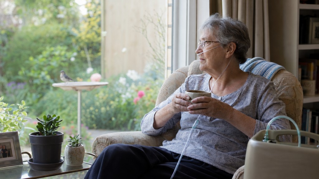 Older woman using oxygen watches a bird in her garden while holding a cup at home.