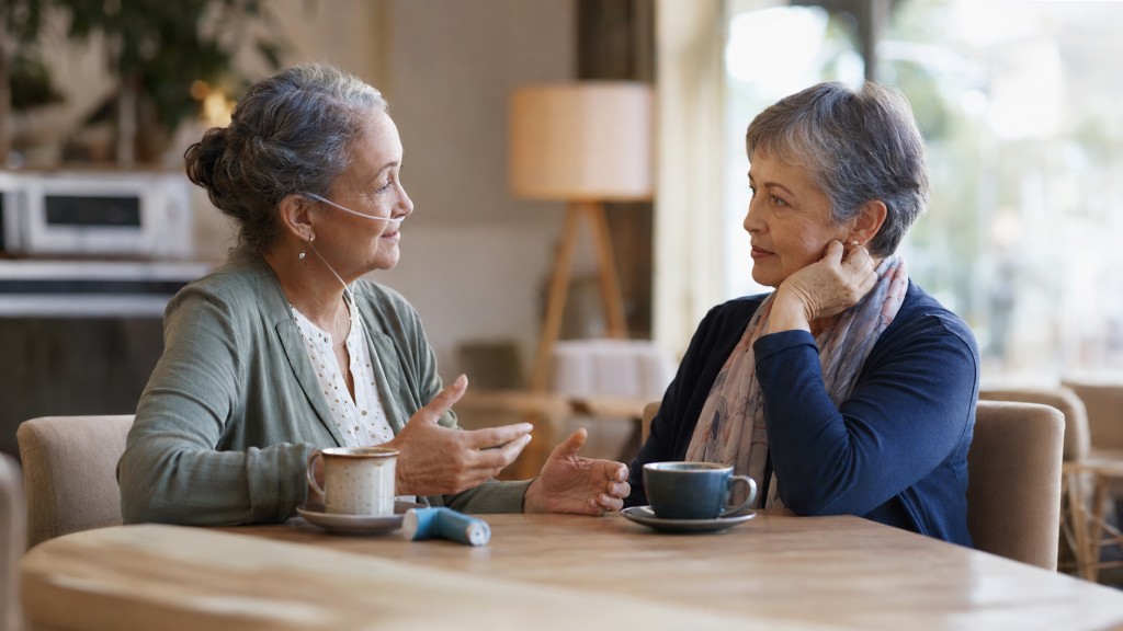 Two older women having a supportive conversation over coffee, one wearing oxygen tubing
